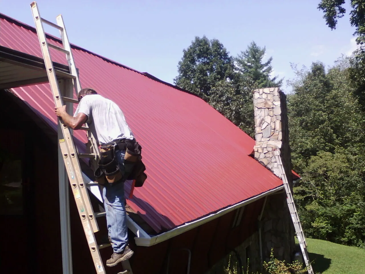 Roofer on a ladder beside a red metal roof and stone chimney — Soffit & Fascia Repair by Roofing Brothers in Saratoga Springs
